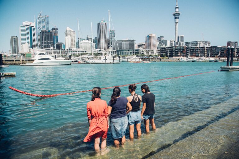 Four female friends standing on the concrete steps down to the marina at North Wharf with their legs in the water and looking towards the city skyscrapers in Auckland